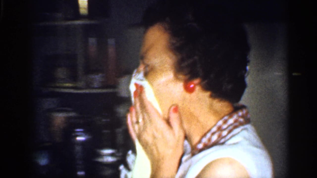 An adult woman wearing a white top with a red-and-white checkered collar wipes her face with a paper towel while standing near shelves in a private kitchen interior.