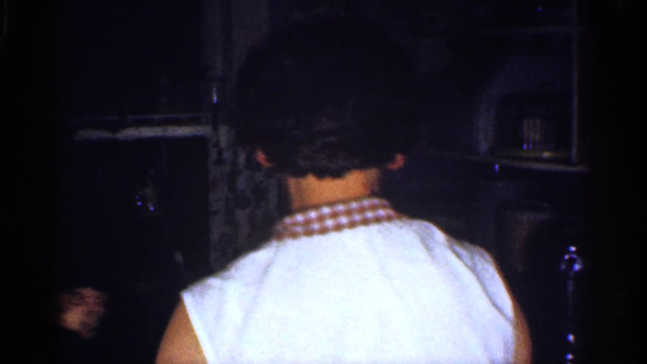 Woman in a white sleeveless shirt seen from behind in a dark kitchen.