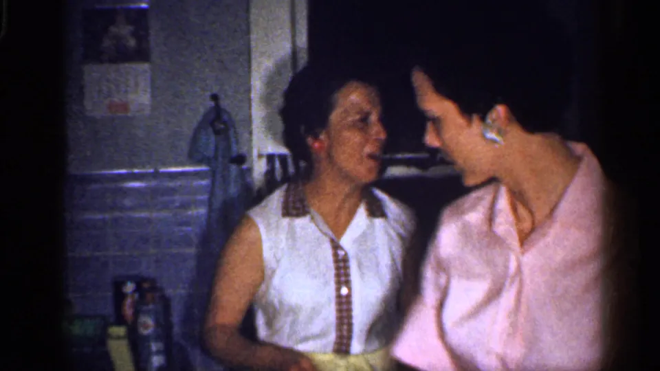 Two adult women stand close together in a 1959 Florida kitchen, one in a white sleeveless shirt and one in a pink shirt.