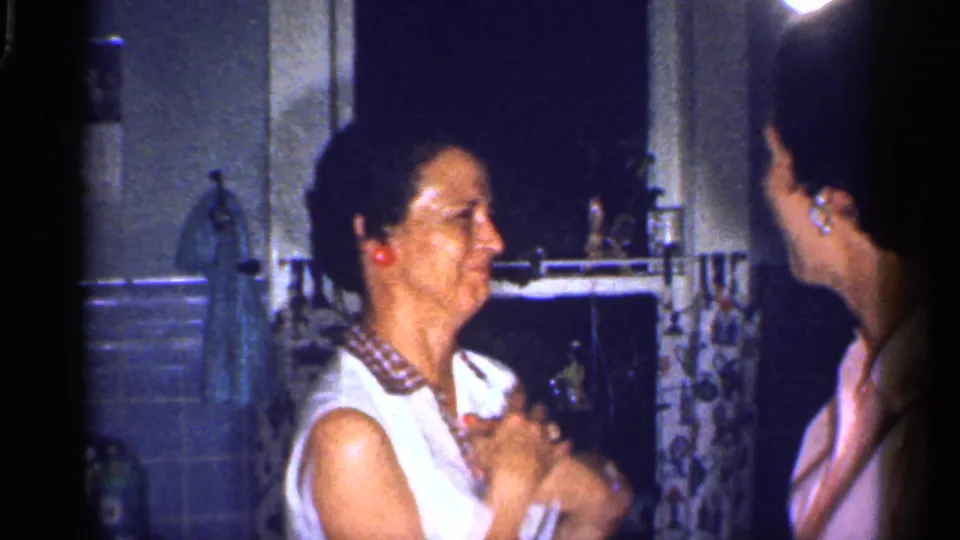 The woman in a white sleeveless shirt smiles toward another woman in a pink shirt in the 1959 Florida kitchen scene.