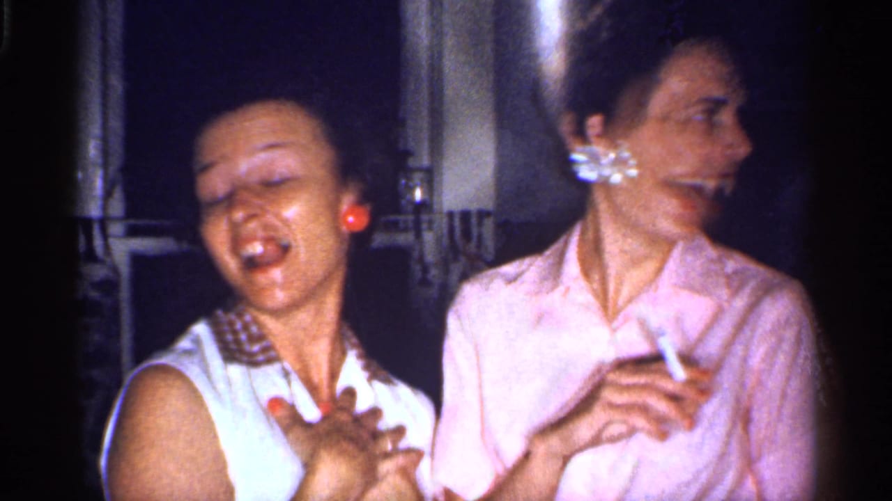 Two women indoors laugh and smoke in dim light.
