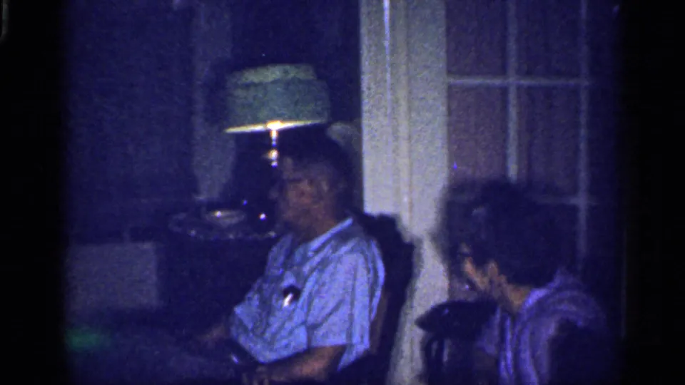 In a dim 1959 Florida interior, a man in glasses sits beside a woman near a glowing table lamp and paneled door.