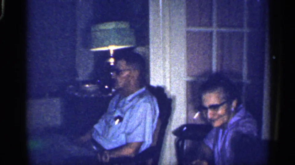 A man in a light shirt and glasses sits near a woman in a dim Florida home-movie interior, with a lamp and door behind them.