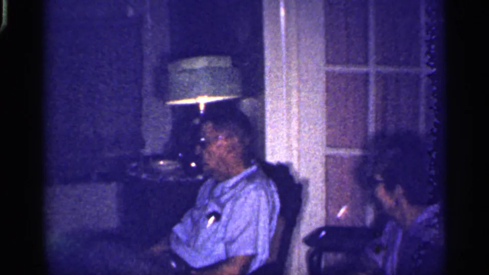 A man in glasses looks toward the camera as a woman sits beside him in the dim 1959 Florida interior near a lit lamp.