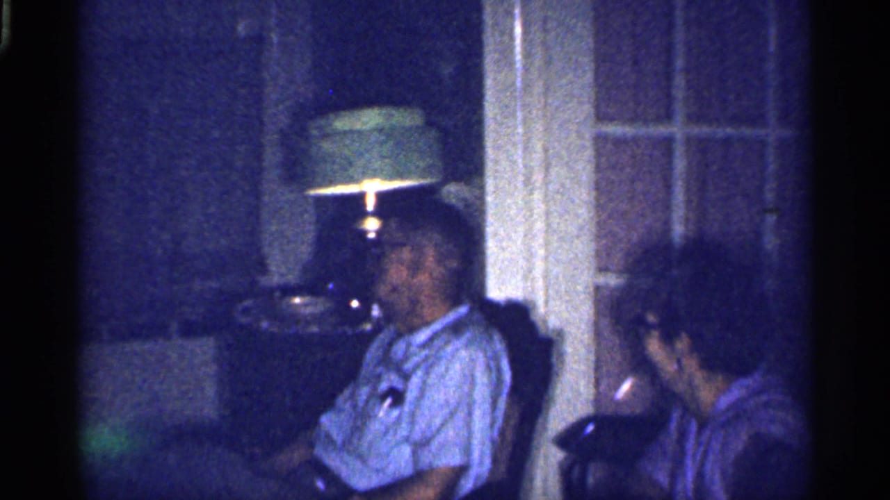 Elderly man and woman sit beside a lamp in a dim living room.
