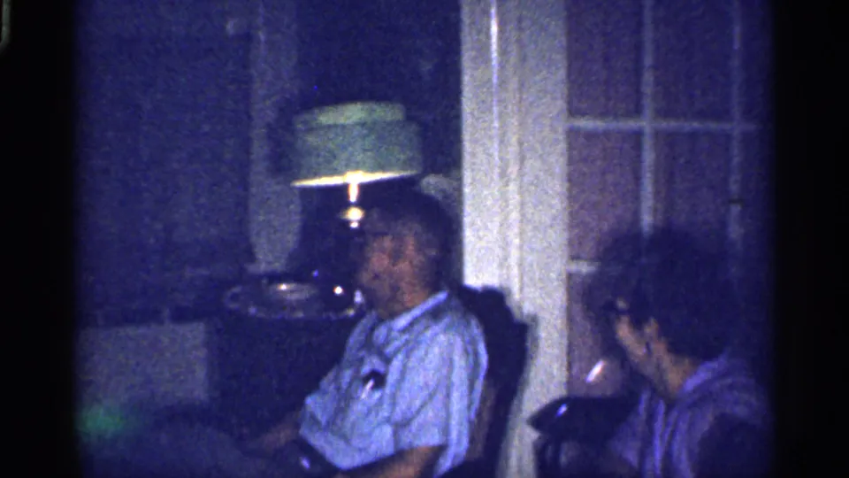 1959 Florida home-movie frame of an elderly man in glasses seated near an elderly woman in a dim private living room.