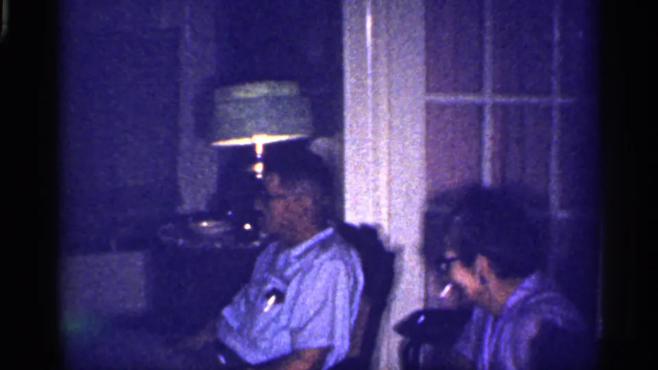1959 Florida living-room footage shows an elderly man in a light shirt seated beside an elderly woman near a glowing lamp.