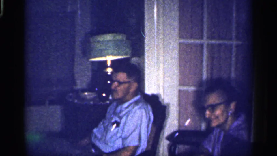In a 1959 Florida private living room, an elderly woman smiles toward the camera beside a seated man in glasses.