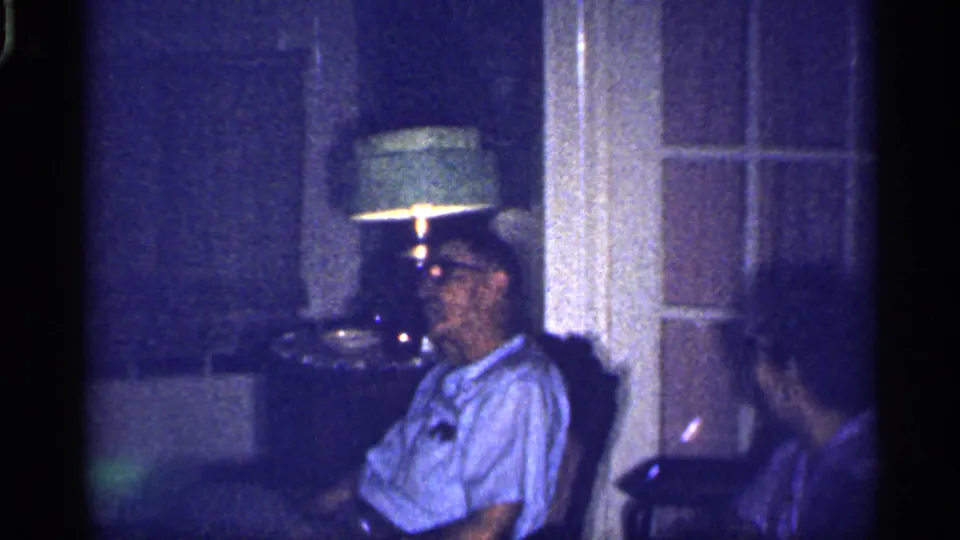 1959 Florida home-movie frame with an elderly man seated by a lamp as an elderly woman turns in a dim living room.