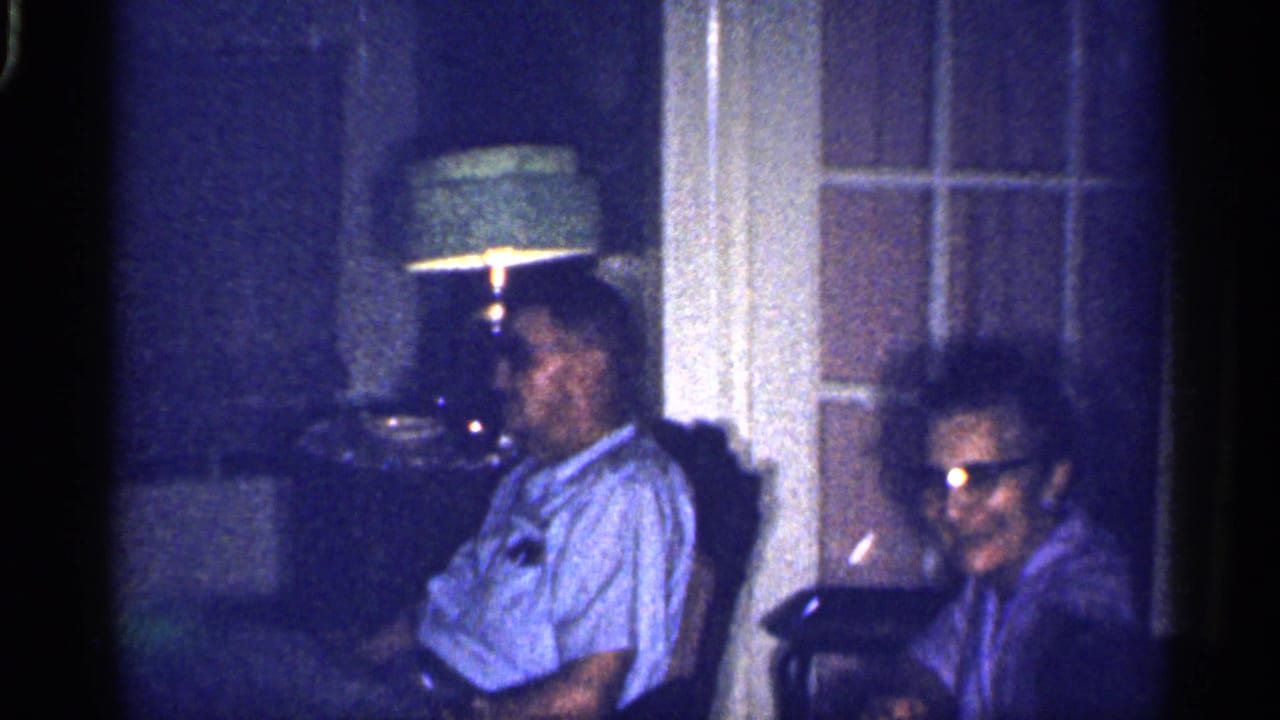 1959 Florida home-movie frame of two adults seated in a dim living room beside a green-shaded lamp and windowed door.