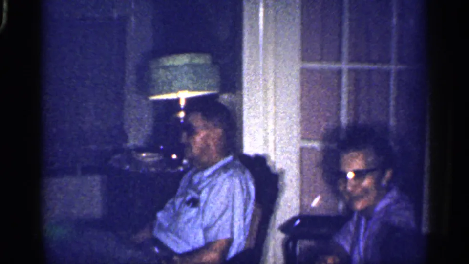 1959 Florida home-movie frame of two adults seated in a dim living room beside a green-shaded lamp and windowed door.