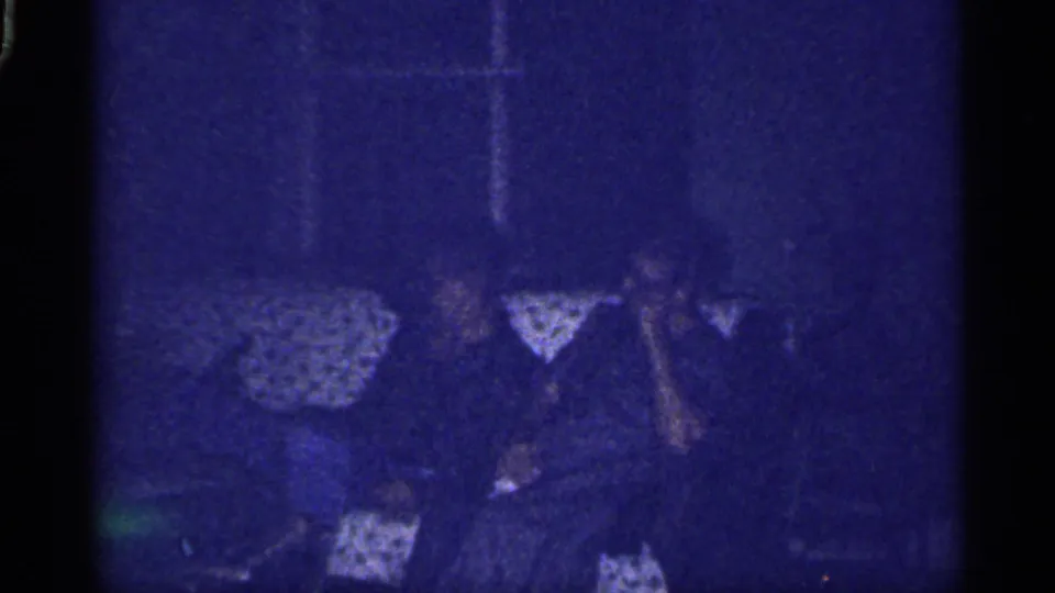1959 Florida home-movie frame of family members, including young figures, seated on a patterned couch in a dim private living room.
