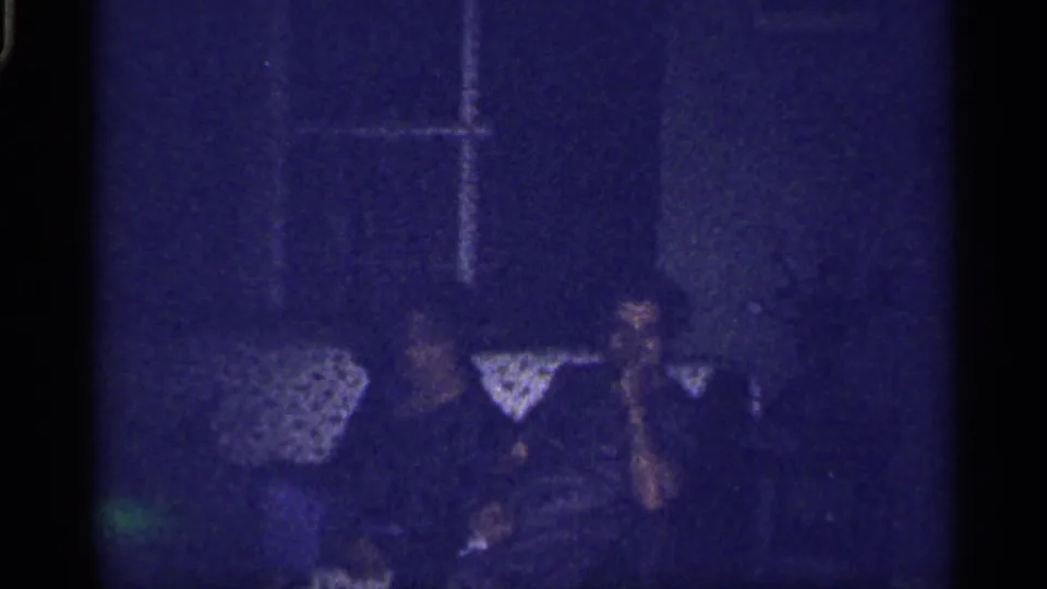 1959 Florida home-movie frame of adults seated close together in a low-lit living room, with a lampshade and window frame visible.