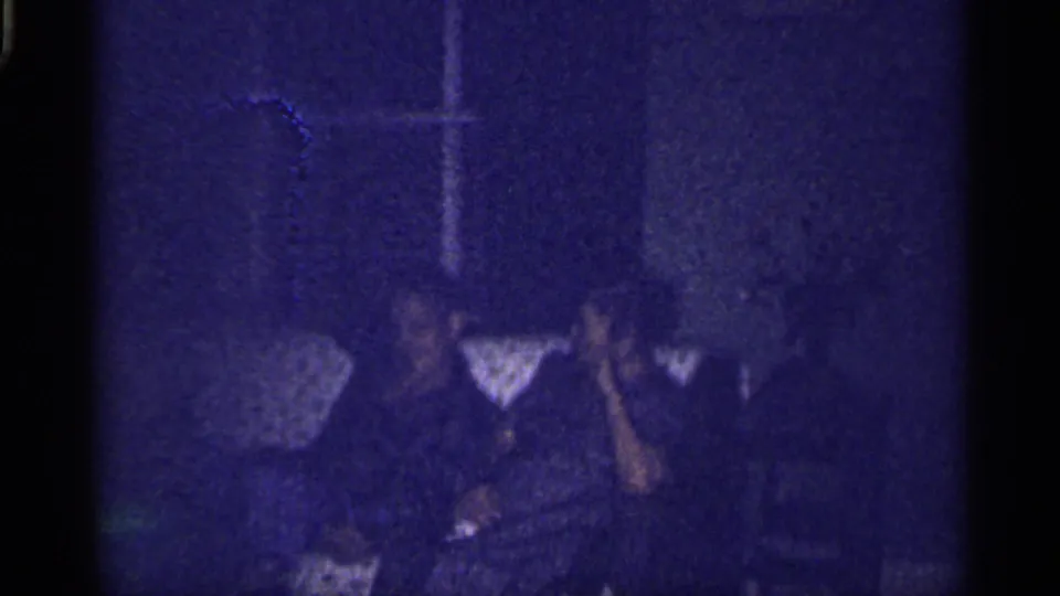 1959 Florida home-movie frame showing a grainy, blue-purple living-room view with seated adults and domestic furnishings.