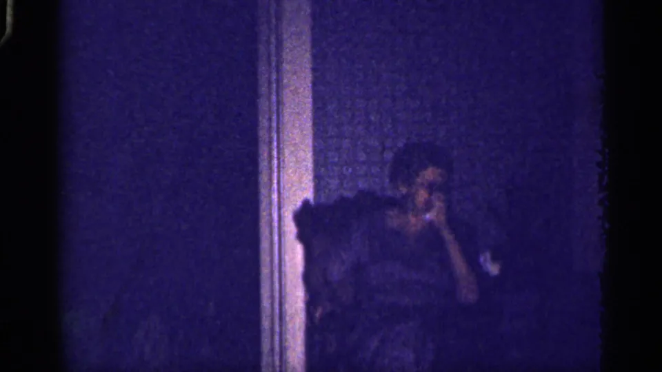 1959 Florida home-movie frame showing an adult seated in a dark living-room interior near a vertical wall or doorway edge.