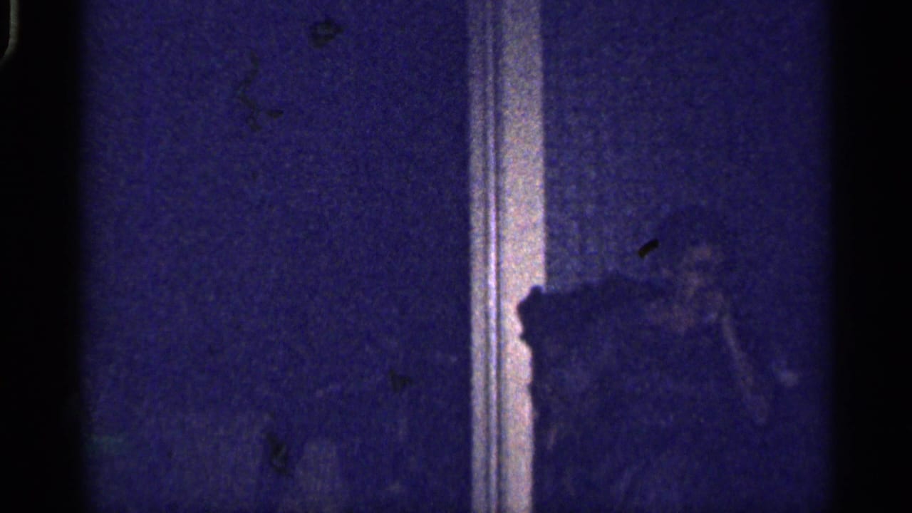 Dim dark-room frame with a barely visible seated person near a doorway or wall trim.