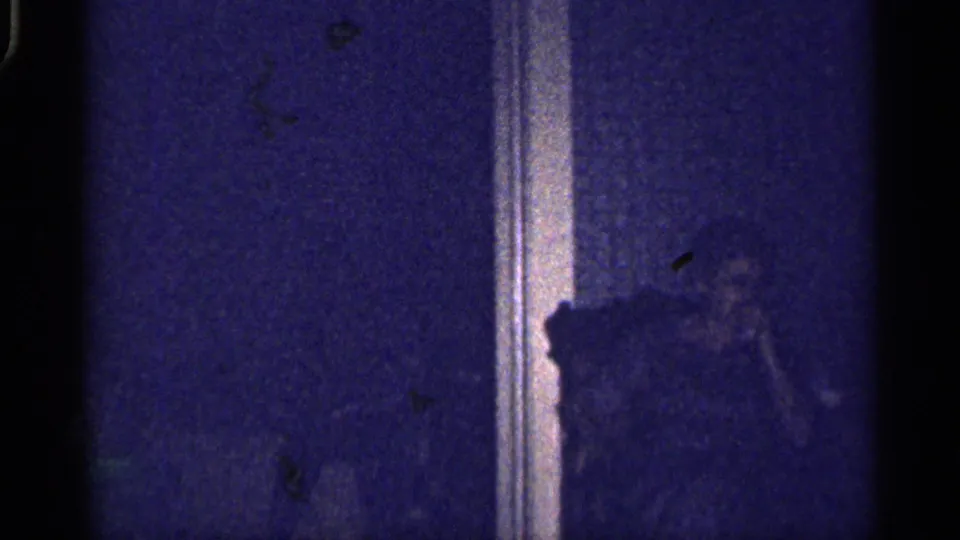 Low-light 1959 Florida home-movie frame with a shadowed seated person in a private room near vertical trim.