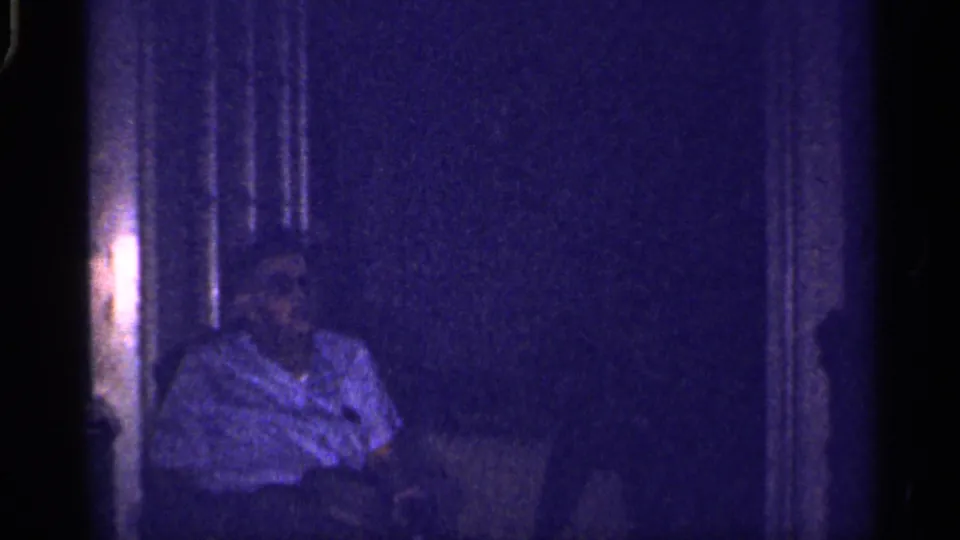 A seated adult in a light short-sleeve shirt appears in a dim Florida home interior, softened by grain and low exposure.