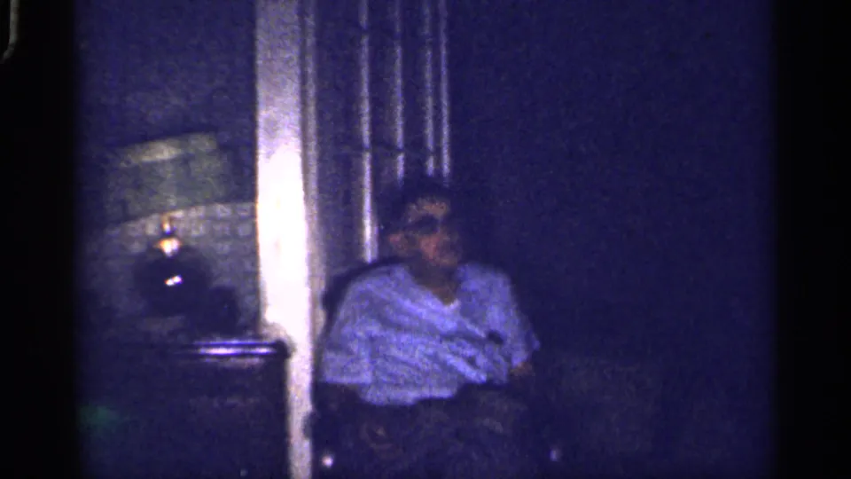 1959 Florida home-movie frame showing a seated adult near a glowing table lamp, patterned wall covering, and dark furniture.