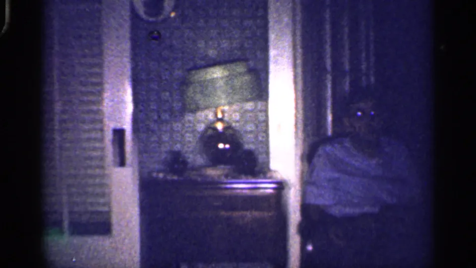 Wider 1959 Florida interior view with a lit lamp on a table, patterned wall, shuttered panels, and a seated family member at right.