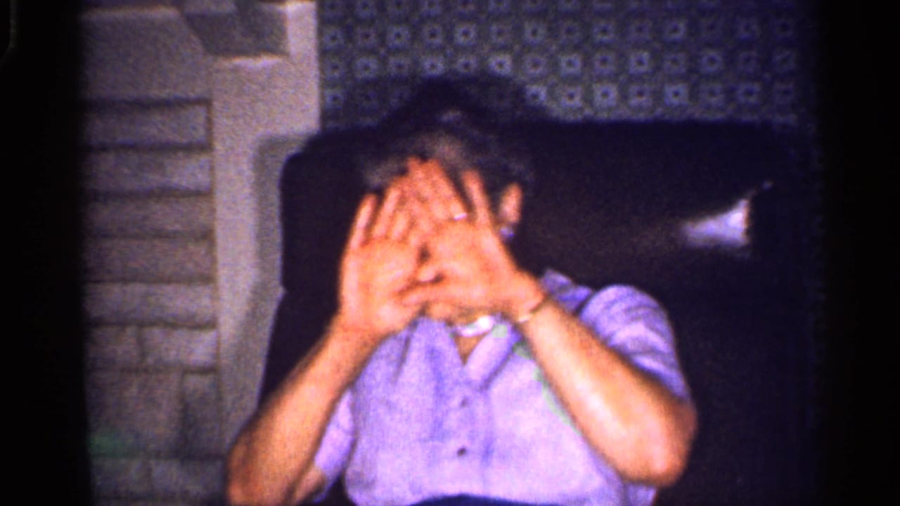 Older woman seated indoors with both hands covering her face.