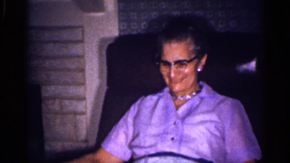 An older woman wearing glasses and a light purple blouse sits in a private Florida interior beside a stone fireplace.