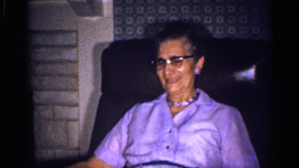 The older woman smiles while seated indoors, with patterned wallpaper and a stone fireplace visible in the 1959 Florida home movie.