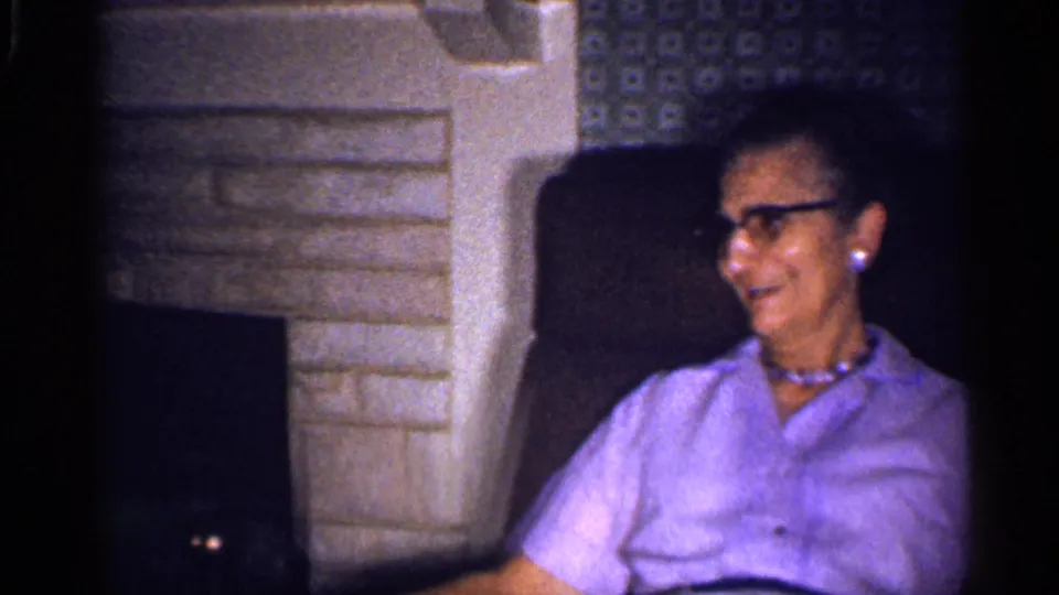 The woman turns to the side and appears to smile or talk from a dark couch in a private Florida living-room interior.