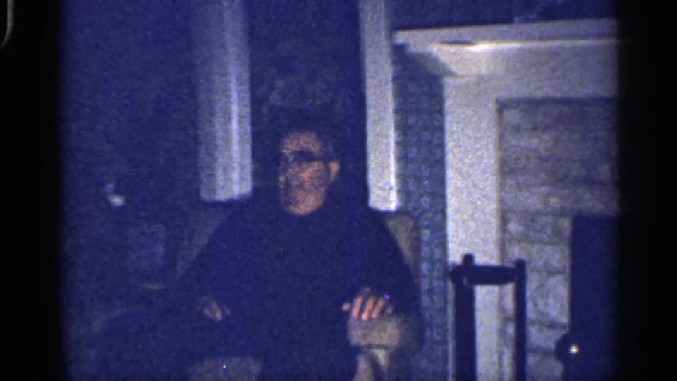 1959 Florida interior frame with an adult man in glasses seated in a chair next to a stone fireplace.