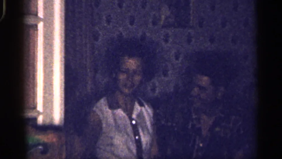 Florida-labeled 1959 8mm frame showing two adults in a dim private interior, one in a light blouse beside a darker-clothed person.