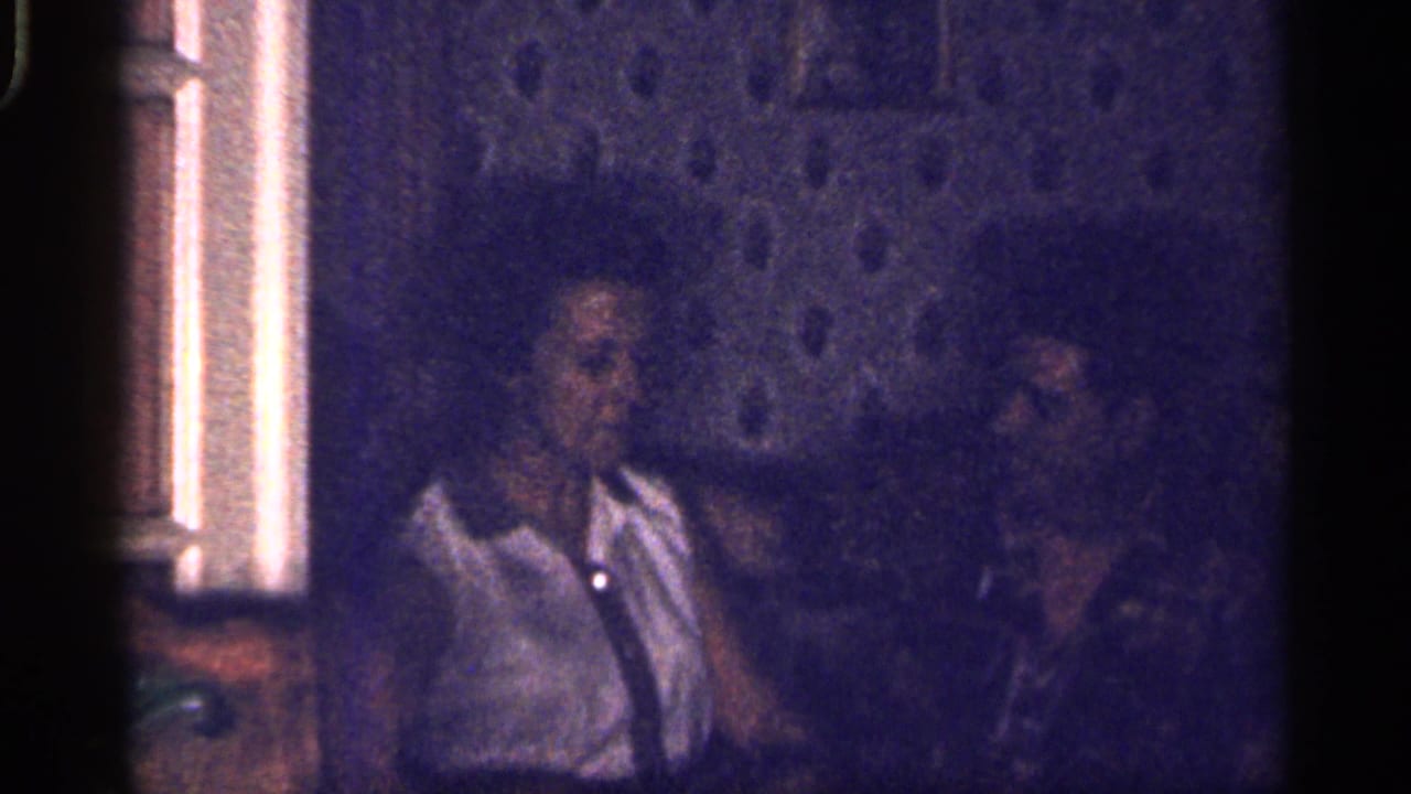 Two adults sit in a dim room beside a textured wall, with the woman in a white collared shirt.