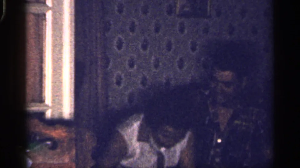 1959 Florida home-movie frame with the woman leaning down and the man seated to the right against a textured wall.