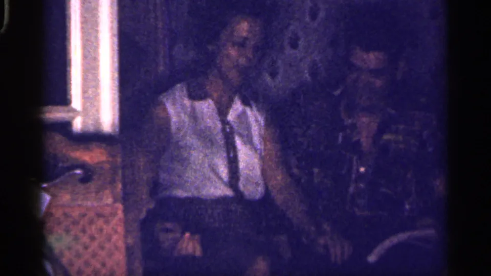 1959 Florida 8mm frame showing two adults seated indoors, appearing relaxed as they face the camera in low light.