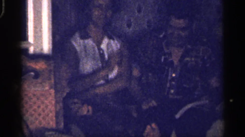 1959 Florida home-movie frame showing adults seated in a dark living room near a curtained window, with soft focus and heavy grain.