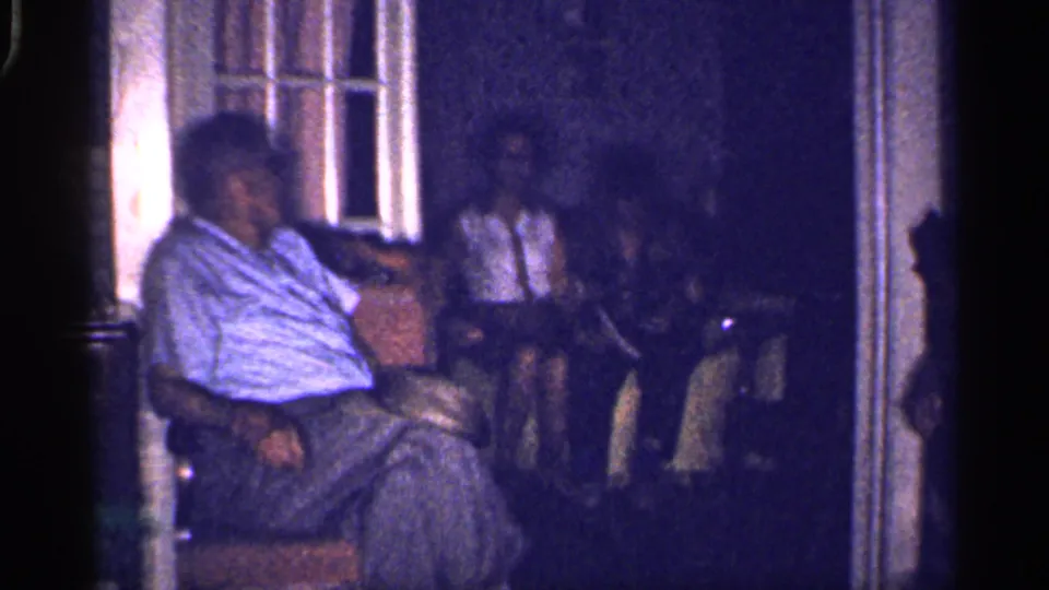 Older man in a striped shirt sits in the foreground of a dim 1959 Florida living room while other adults sit behind him.