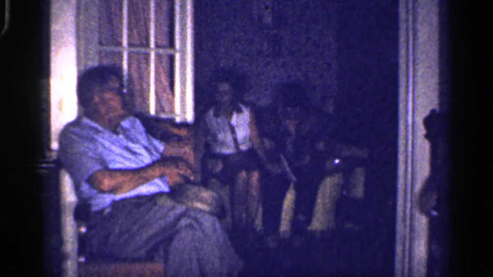 In a 1959 Florida living-room scene, an older man sits in front while a woman behind him rests with bare feet visible.
