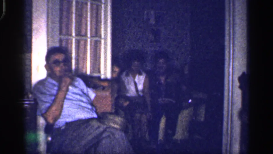 1959 Florida 8mm frame of a man in a striped shirt facing the camera as a barefoot woman and other adults sit in shadow behind him.