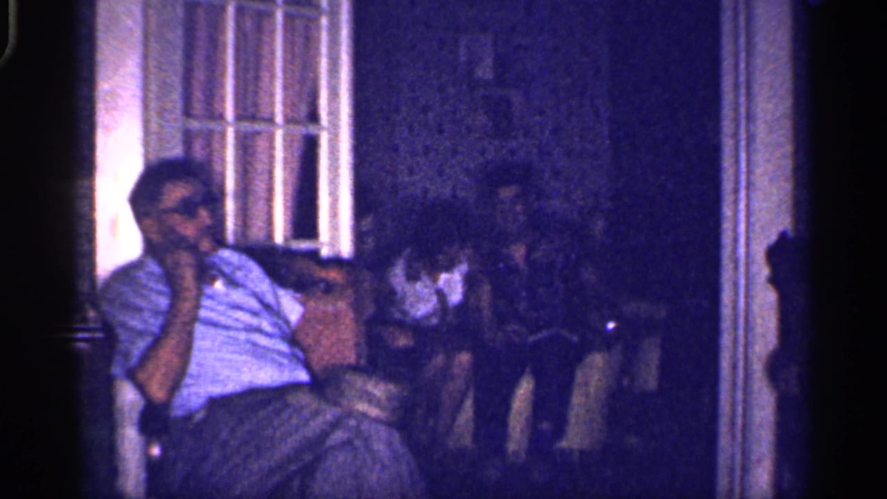 Man in glasses sits in a dim room near a window with other figures in the background.