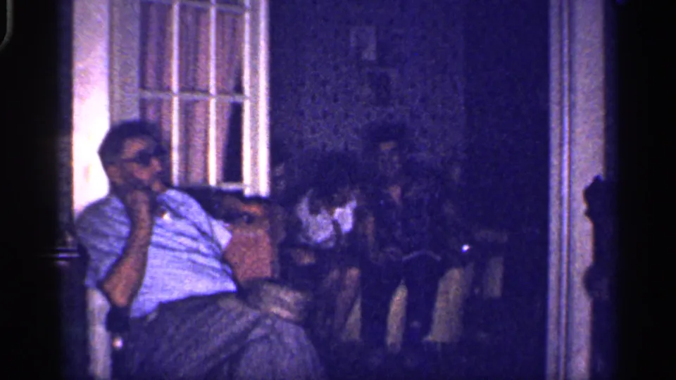 1959 Florida home-movie frame showing a seated man in glasses near a multi-pane window while others sit in the dim interior behind him.