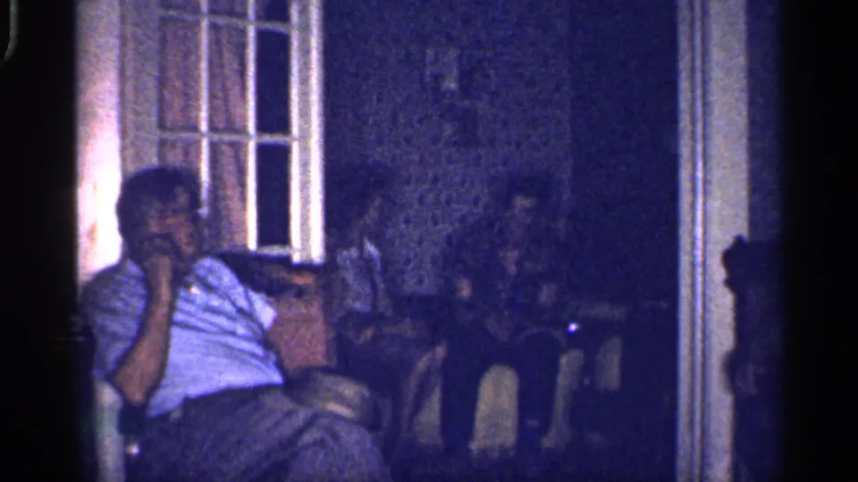 A seated man in a light shirt and glasses rests his hand near his face during a dim 1959 Florida family gathering indoors.