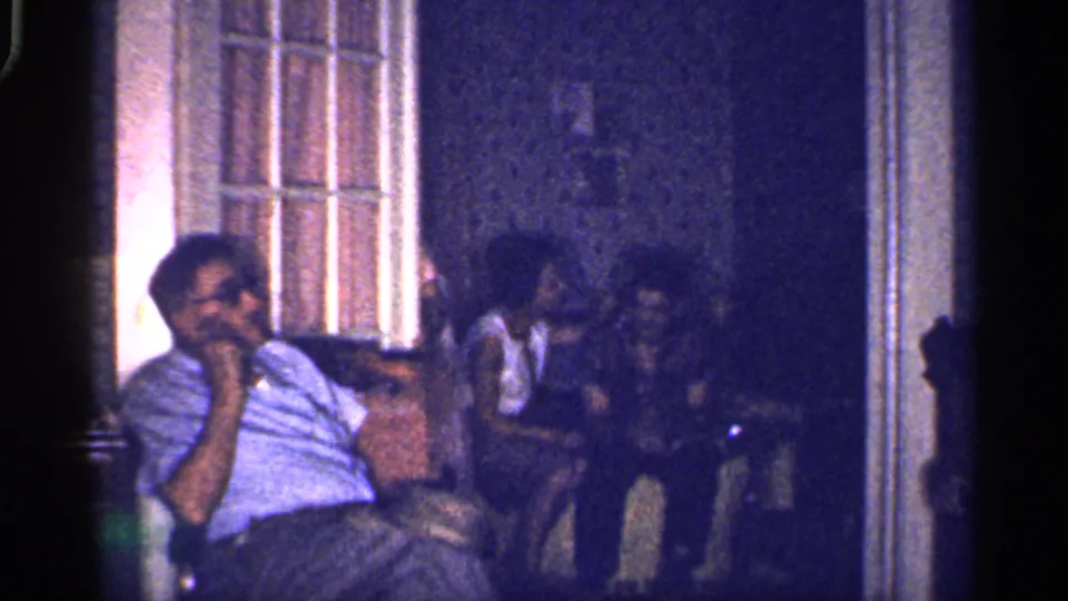 1959 Florida home-movie still of a seated man in glasses with shadowed family members and chairs visible in the background.