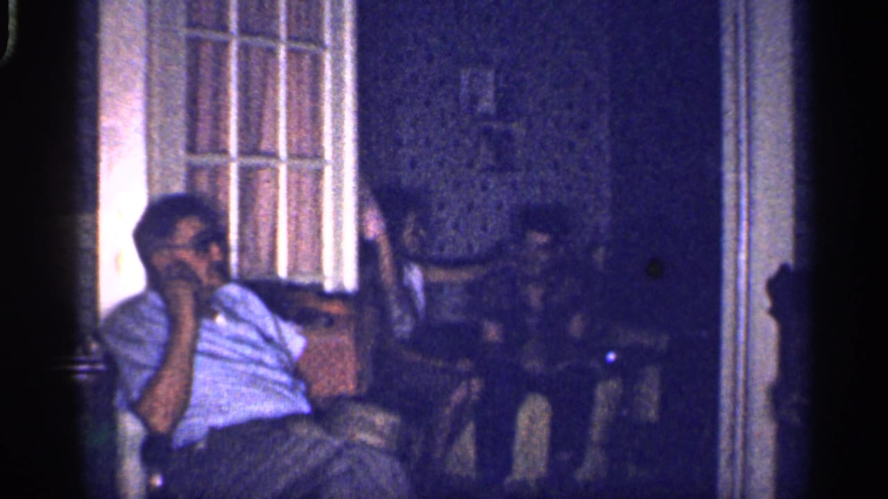 1959 Florida home-movie frame showing adults seated indoors, including a man in glasses near a tall window in a dim private room.