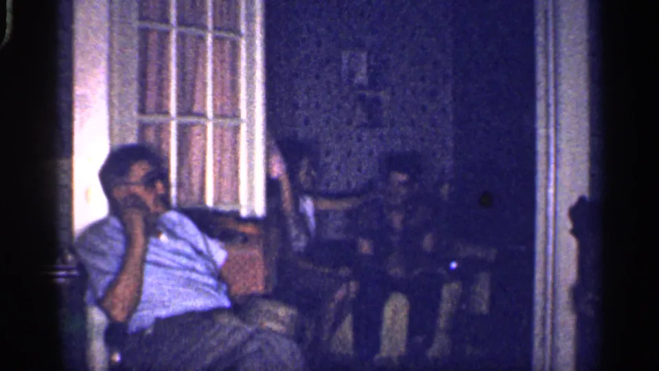1959 Florida home-movie frame showing adults seated indoors, including a man in glasses near a tall window in a dim private room.