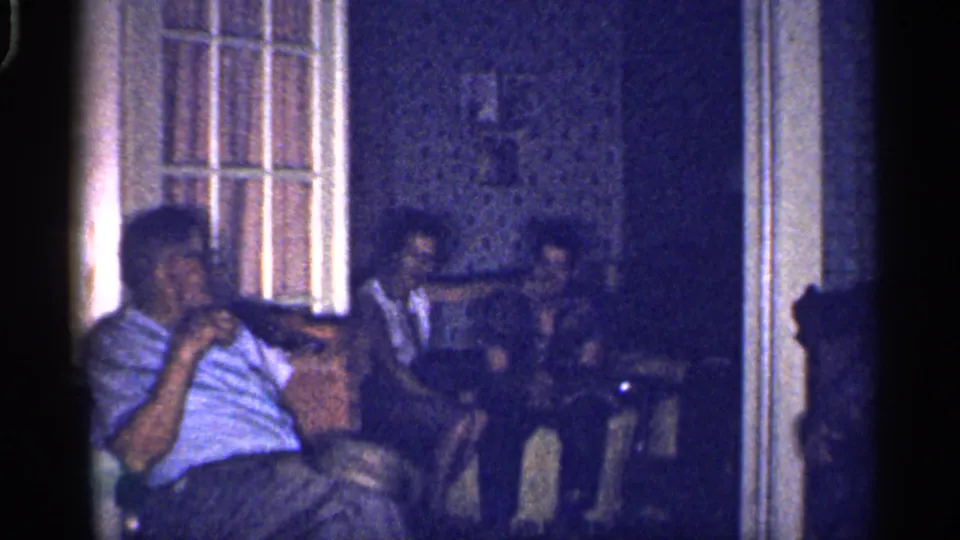 1959 Florida 8mm frame with a seated man in the foreground and several adults grouped in a low-lit private living room.