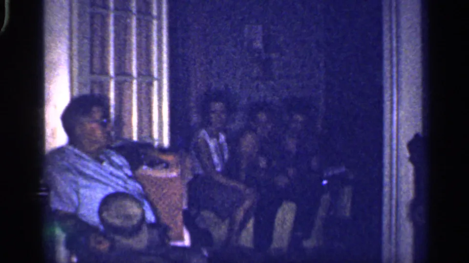 Blue-tinted 1959 Florida home-movie frame showing adults seated indoors, partly obscured by low exposure and 8mm grain.