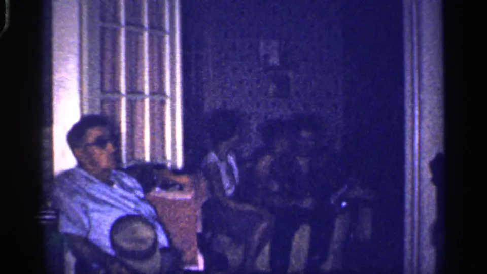 1959 Florida frame of a quiet indoor family gathering, with adults seated in shadow beside a tall window or door.