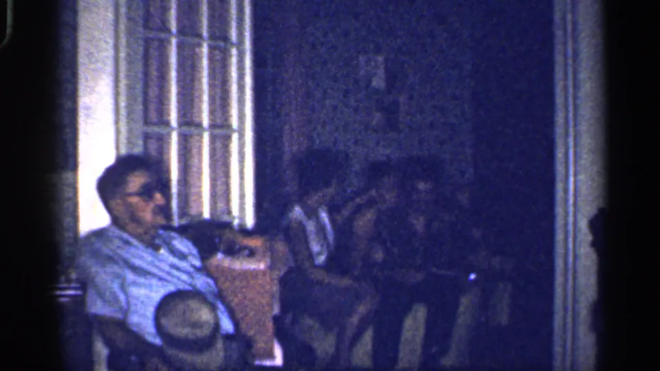 1959 Florida 8mm frame showing adults seated indoors, with a man in sunglasses in the foreground and others in shadow nearby.