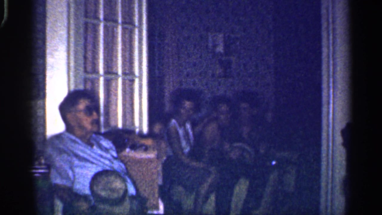 Seated man in sunglasses faces several adults in a dark interior room.