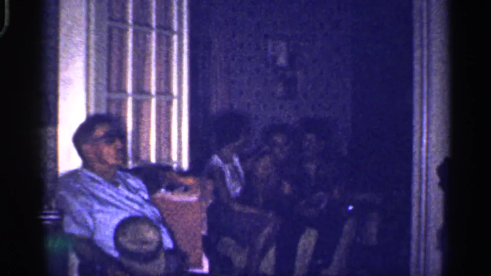 1959 Florida 8mm frame showing adults gathered in a dim private room, with the foreground man lit more clearly than the others.