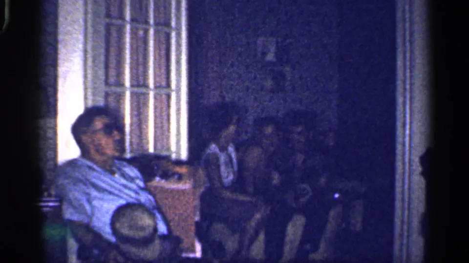 1959 Florida home-movie frame with adults seated in a private interior, including a man in sunglasses near a bright paned opening.